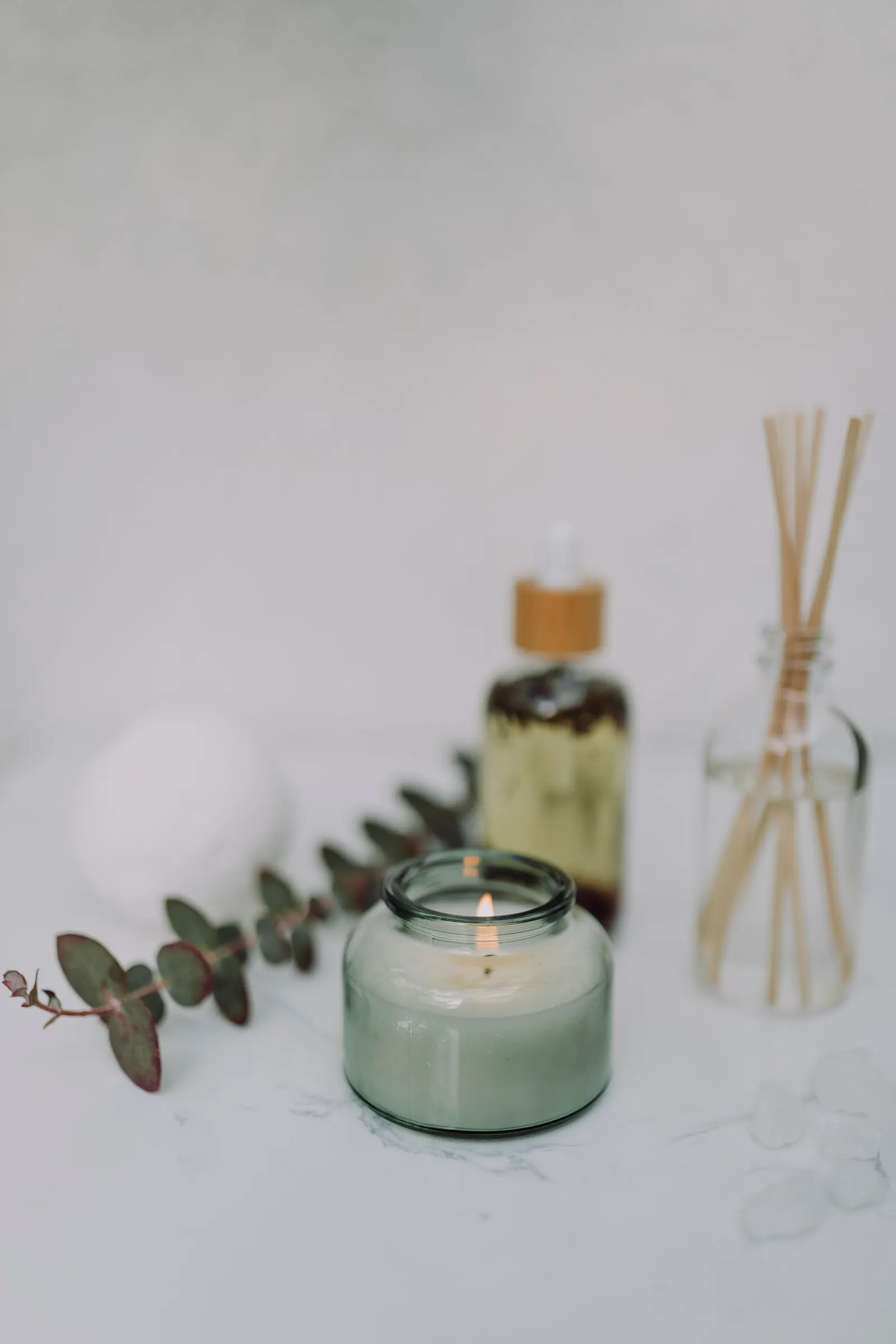 Lit scented candle with diffuser and eucalyptus leaves on white marble